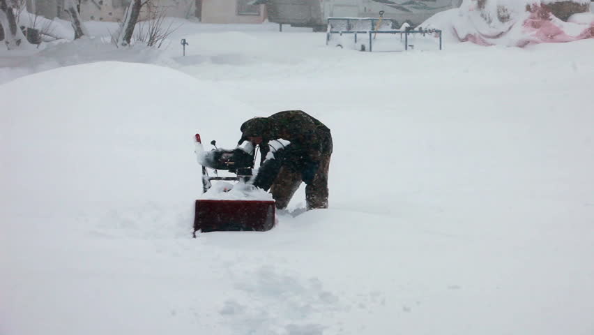Man tries to start a snow blower stuck in deep snow blizzard. Snow blowing in strong wind in sub freezing weather. Winter time hazard and climate nature.