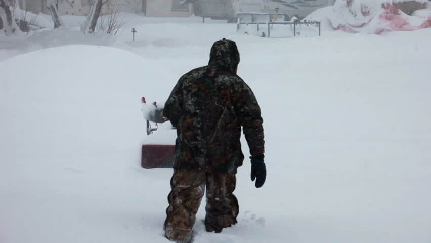 Man walks to snow blower stuck in deep snow blizzard. Snow blowing in strong wind in sub freezing weather. Winter time hazard and climate nature.