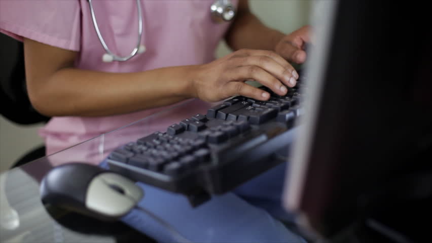 A pretty young African American nurse or nursing student working at her computer stops typing and smiles at the camera. 