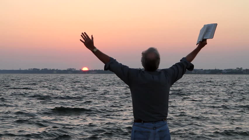 Man holds his arms up with a bible in hand as he prays in front of a body of water at sunset.