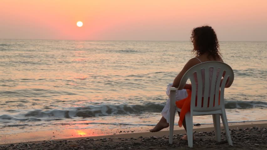 curly-headed woman sits on white plastic chair alone on beach faced to sea at sunset