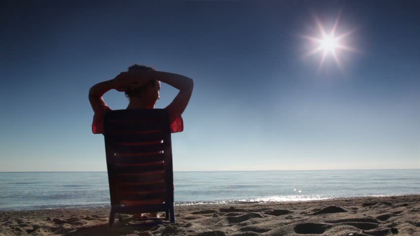 Woman sitting on deckchair and tans holding hand behind head, on beach at sunny day, backview