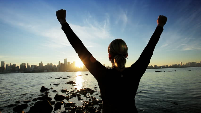 Female in silhouette saluting the challenge of a new day with a city skyline backdrop at sunrise