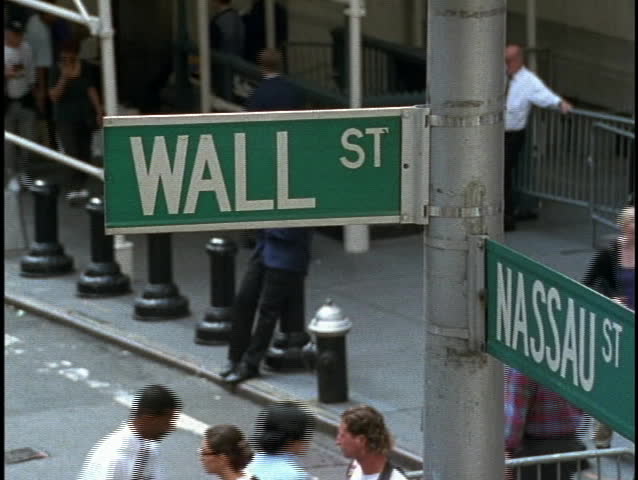 Crowds of people walk on the corner below the street signs of Wall and Nassau street in New York City.