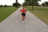 A pretty brunette with remarkable muscle definition jogging outdoors on an asphalt jogging track.  Tracking shot, full-length, front view.