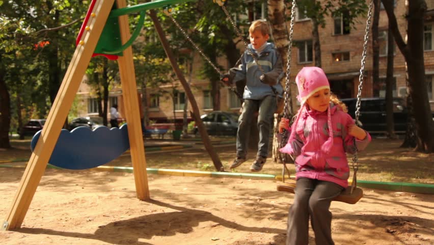Brother and little sister sway on swing at playground, sunny autumn day