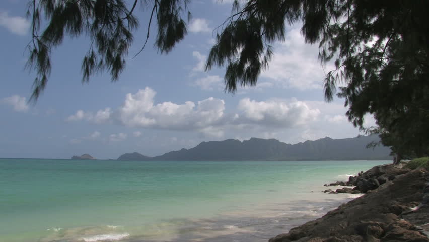 Tropical beach on Oahu, Hawaii. Islands off shore in Pacific ocean. Outriggers paddling by distance islands. Waves on white sand with tree limbs overhead. Scenic, relaxing, calm seascape. Pan left.