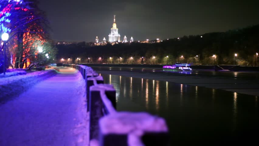 Motor ship decorated with garlands, floats down the river, quay with colorful illumination at winter night, building of Moscow University on background