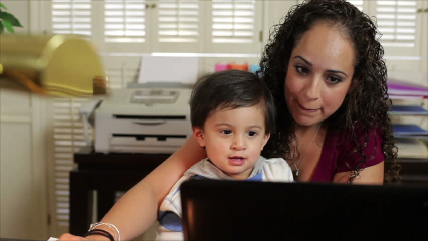 A pretty Hispanic mom in her home office shows her little son something on the laptop.