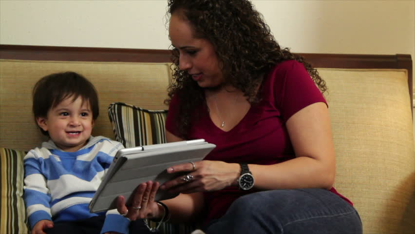 A cute little Hispanic toddler playfully imitates something he sees on the electronic tablet his mother is holding.