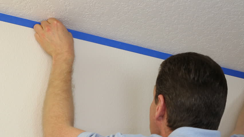 Man preparing to paint a white ceiling by masking off the wall beneath it with blue painter's tape.