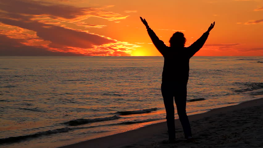 Woman stands on the beach raising her hands into the arm in prayer and praise worship as the sun sets on the ocean.