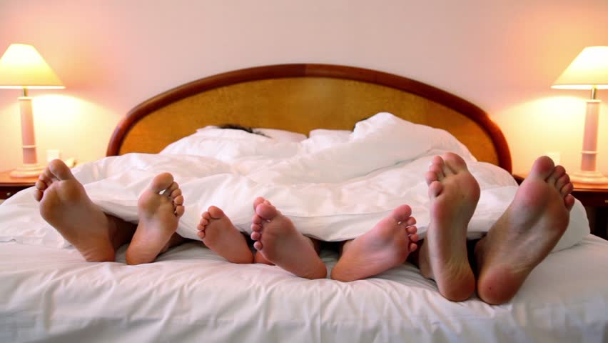 Parents and their kids lay in bed under blanket and move bootless feet, closeup view
