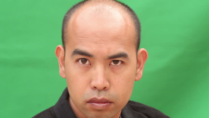 An Asian man in a Japanese Gi draws a Samurai Sword (Katana), and holds it in a threatening high en garde position in front of a green screen background.