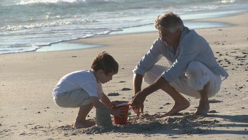 LONG SHOT OF A WOMAN PHOTOGRAPHING A GRANDFATHER AND GRANDSON MAKING SANDCASTLES