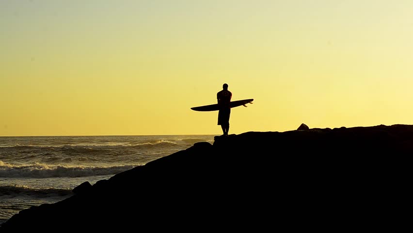 Male surfer holding his surfboard watching the waves at sunset.