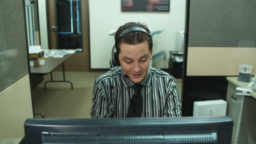 office worker laughs and uses computer while talking on headset; out-of-focus co-worker passes in background (no audio)