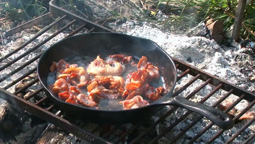 Bacon cooking in a black cast iron skillet over a smoking campfire.
