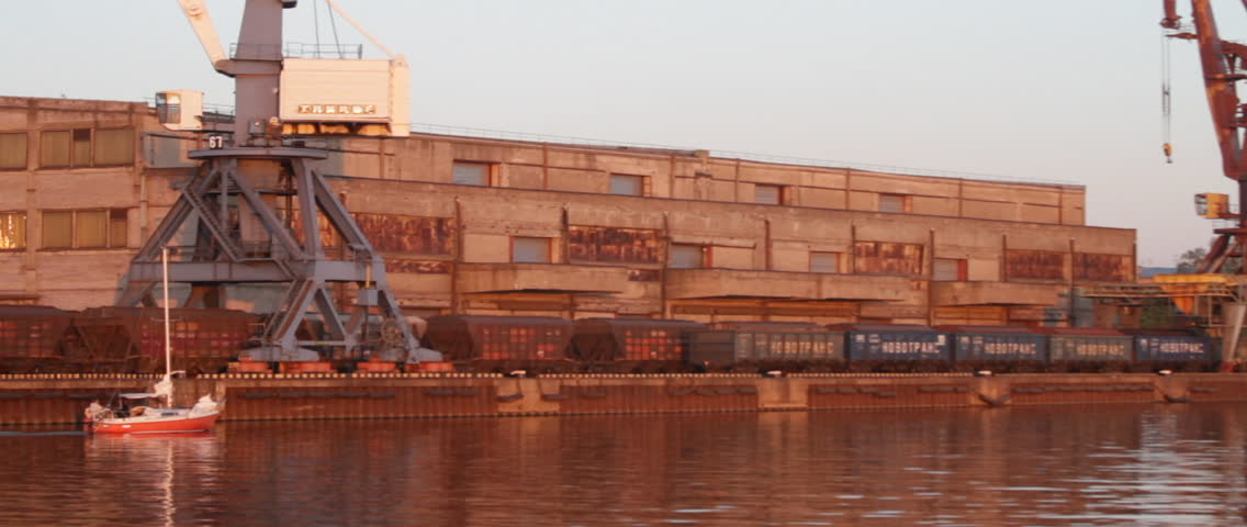 Sailboat moving at the Riga Freeport along the Daugava river, in Riga, Latvia. Port crane, container terminal and storage facility in the background.