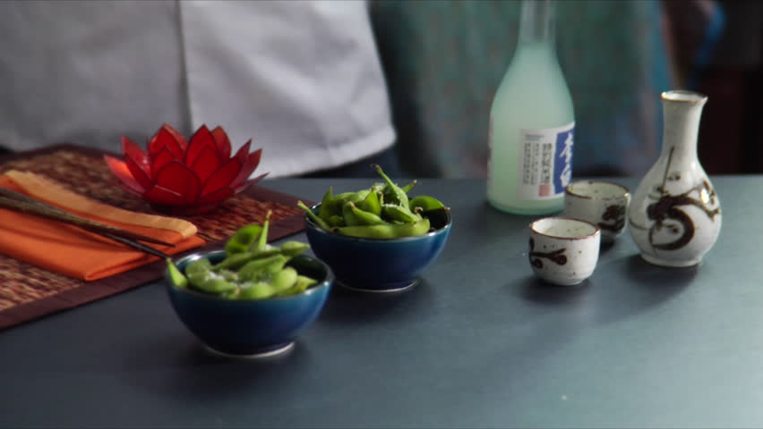 Lock down shot of chef pouring hot sake with bowls of Edamame (soy beans) on the side.