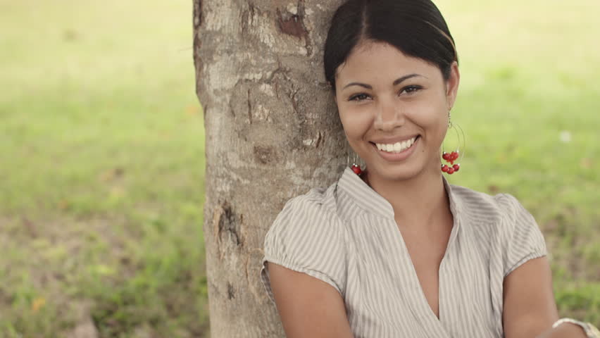 Portrait of happy young latina woman smiling under tree in park. Tilt shot