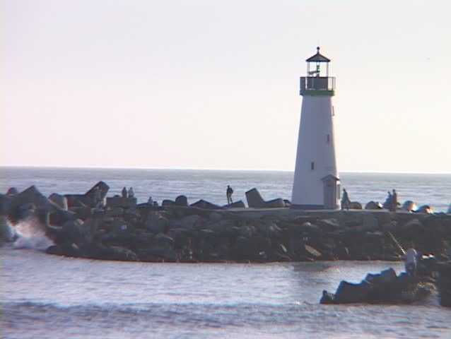 Medium shot of a lighthouse with people observing the water and a gull swooping towards the surf.