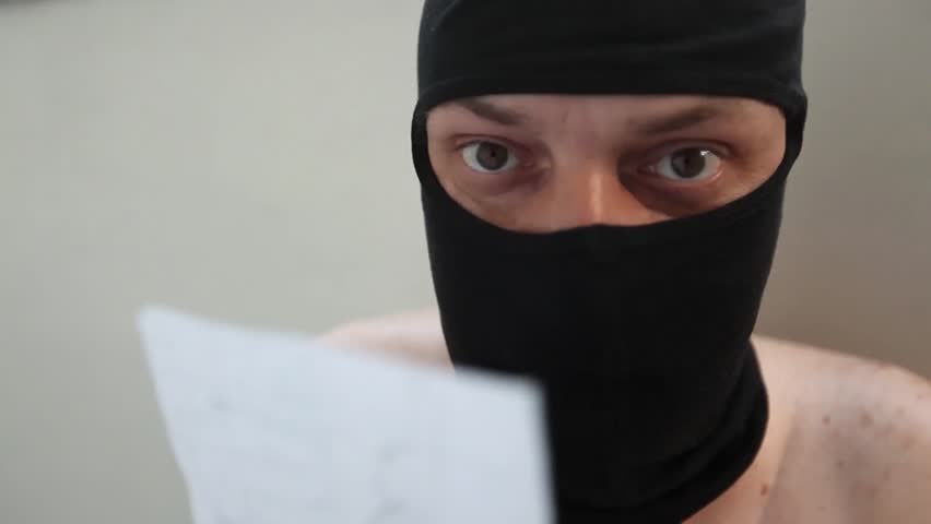 Naked young adult man with black balaclava reads a paper.
