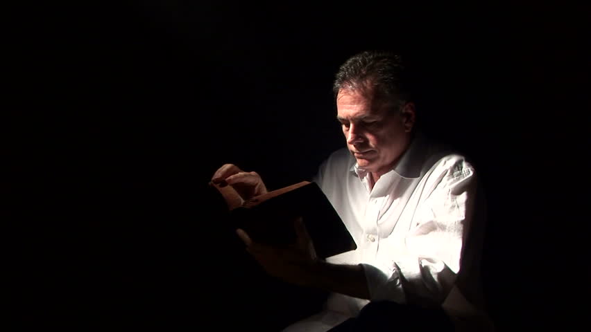 A man spot lighted by a fog filled beam of light, reading a bible and praying. 