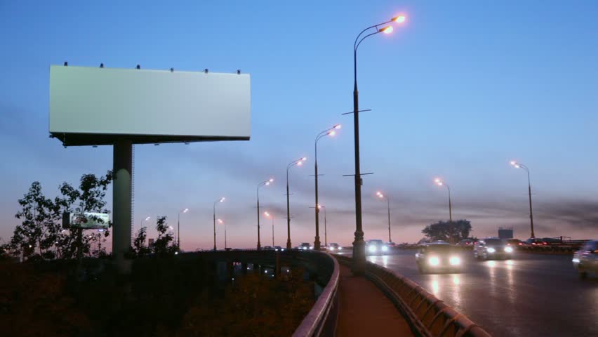 Empty advertising pillar on sidelines of highway with traffic at evening