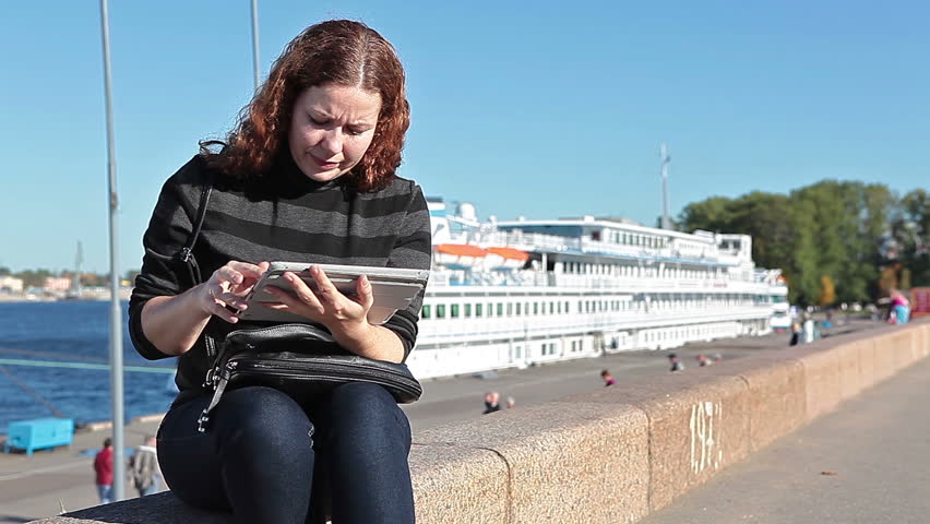 Young beautiful Caucasian woman with tablet computer sitting on city embankment