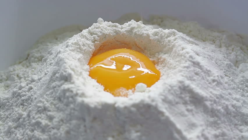 Fresh yellow egg being broken and  dropped down into white baking flour