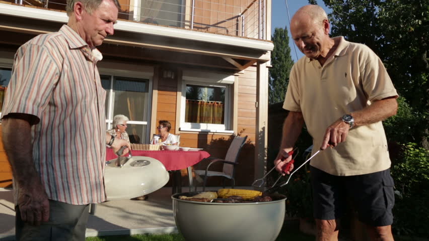 senior men cooking on kettle barbecue women sitting on terrace in garden