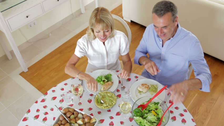 Senior couple seated around dining table serving one another from above.