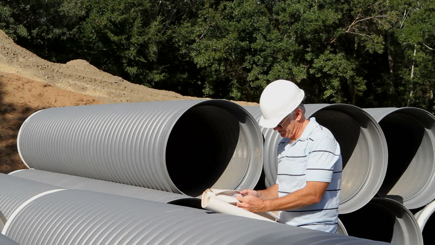 Civil engineer stands by a stack of drainage pipes and checks the specification on the blueprints at an excavation site.