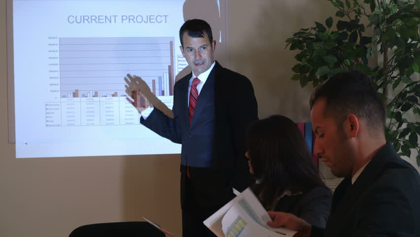 A projector shines a graph on the screen and on a businessman explaining the information. 