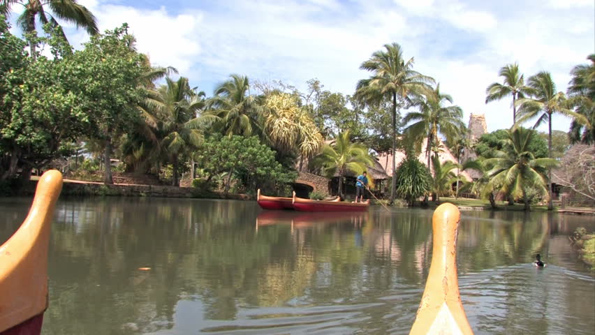 Polynesian canoes in a tropical pool in Oahu, Hawaii. Passing. Palm trees and tropical huts. Polynesian Cultural Center, Hawaii number one tourist attraction. Show the various islands of Polynesia. 