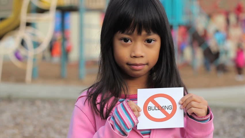 A cute Asian girl holds up a small NO BULLYING sign on the school playground.
