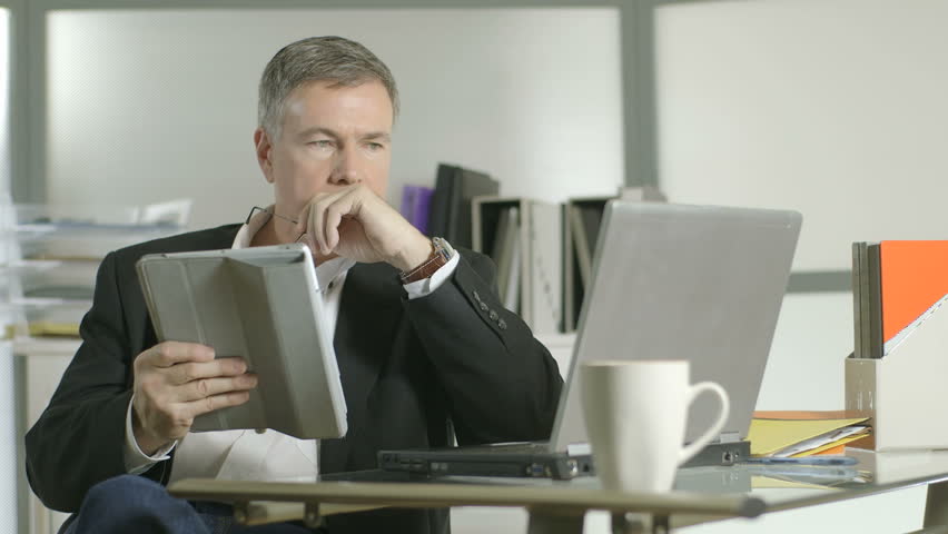 A mature handsome businessman in his office working simultaneously with an electronic tablet pc and a laptop computer.