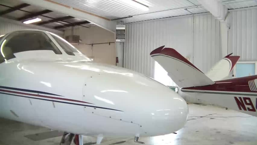 Panning of small jet airplane in hanger from nose to wing with shiny white fuselage glimmering in natural lighting.