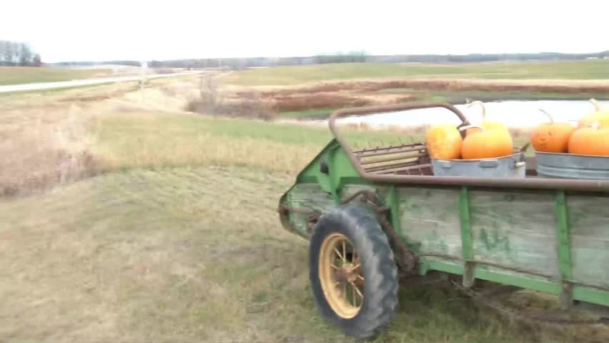 Pumpkins are atop aging green farm tractor trailer during October in rural Minnesota with small lake in background.