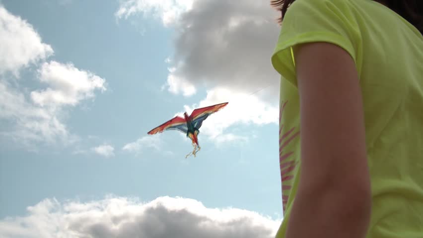 Girl is flying kite on partly cloudy afternoon, shot from lower rear with her maneuvering string in wind, sun shines through at end.