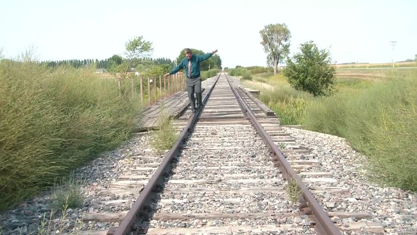 Businessman walks a fine a line down one side of a railroad track, stops to discover something, and resumes past camera, sunlit lighting.