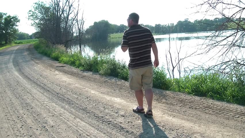 Man drinks beer on a dirt road after camera pans from cornfield to exquisite lake.