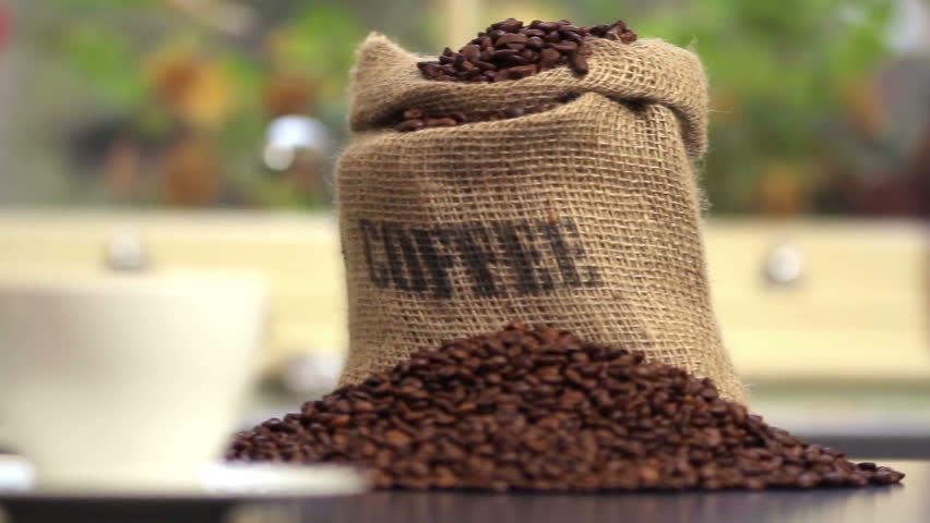 Steaming cup of coffee with coffee beans overflowing out of a hessian coffee bag behind. Camera tacks along kitchen worktop and pulls focus on white cup with garden in the background.