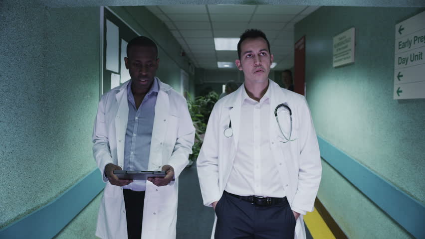 Two male doctors discuss a patient's notes as they walk down a hospital corridor. Other medical staff and patients can be seen in the background. 