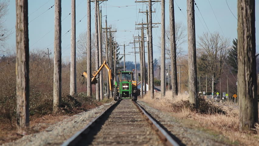 A specialized  machine is used for trimming weeds and plant growth alongside railway tracks/Maintaining Railway Tracks/A long arm and blade trims overgrowth along railway tracks.   