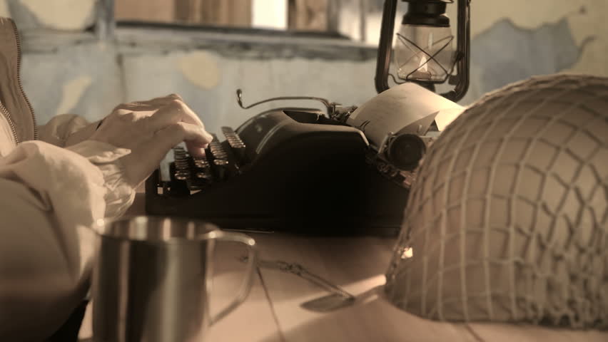 A man who appears to be a soldier sitting at a makeshift table in a bombed out building typing on a manual typewriter.