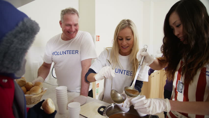 Friendly voluntary workers standing at a soup kitchen serving hatch are handing out cups of hot soup and bread to a waiting line of homeless and needy people. In slow motion