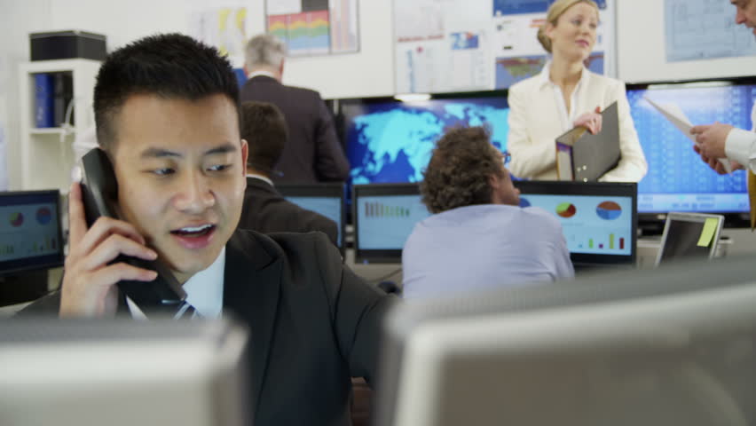 Portrait of a young and ambitious stock market trader working in a busy office filled with computers. The rest of his team are hard at work in the background. 