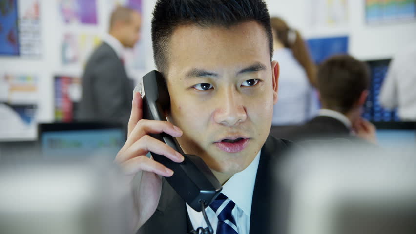Young and ambitious stock market trader is doing a deal over the phone in a busy office filled with computers. The rest of his team are hard at work in the background. 